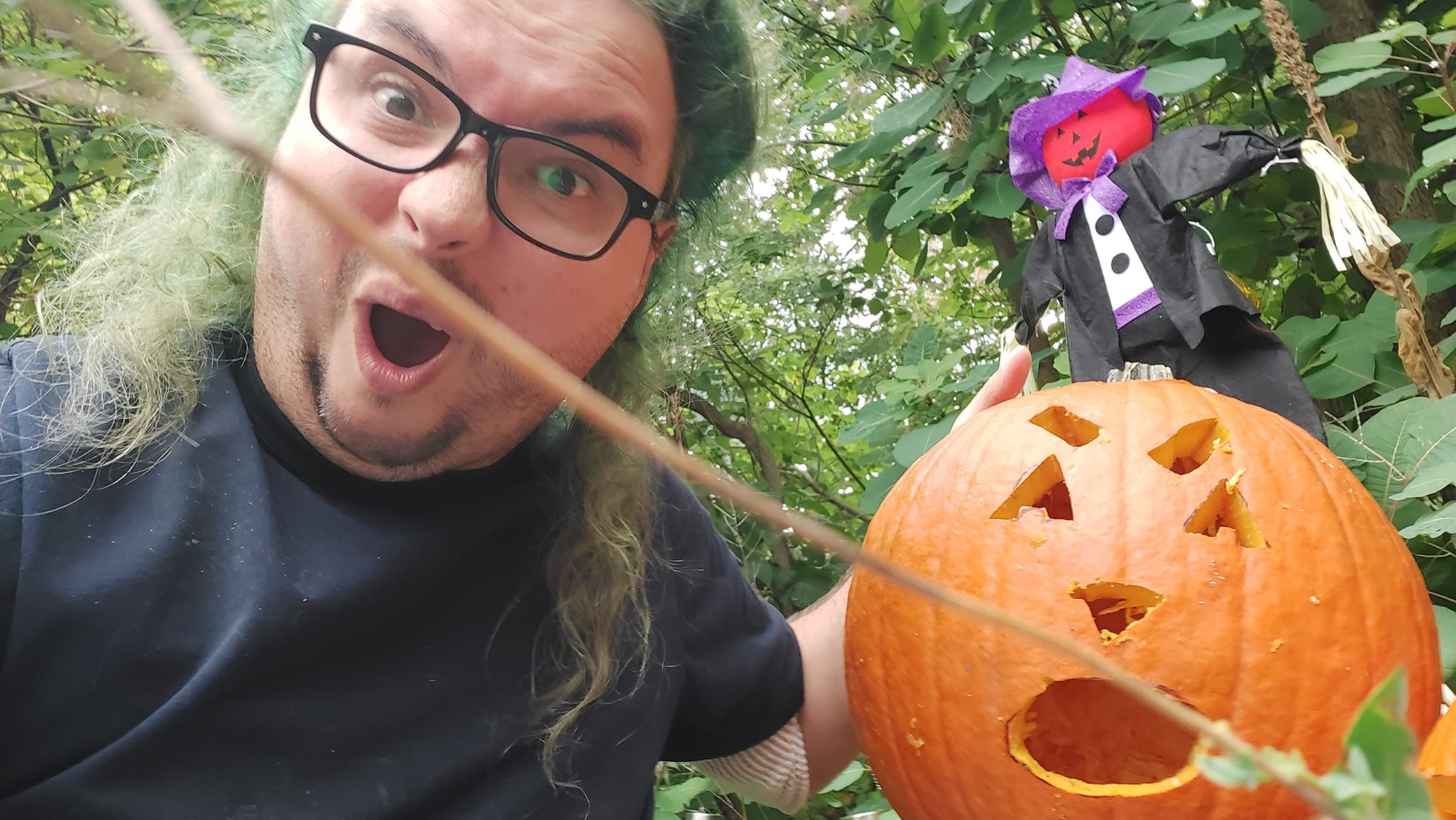 A man with long green hair and no beard has a yelling expression and holds a large punpkin with a suprrised face carved on it